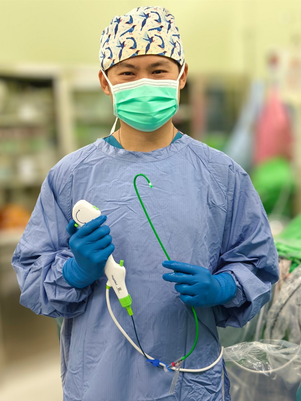 黃韋銘醫師在手術室內，身著藍色手術衣、戴著口罩與印有圖樣的手術帽，雙手各持著一段手術用的內視鏡器械，面向鏡頭