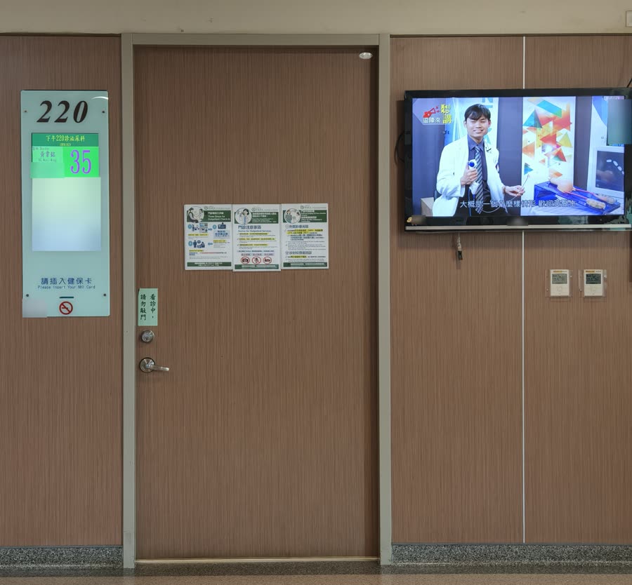 安南醫院門診 220 診的門口：木紋門左邊是寫著 220 的叫號看板，看板下方寫著「請插入健保卡」，門右邊牆上有一台電視
