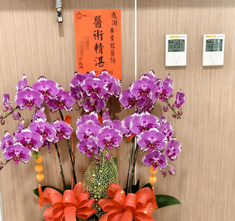 醫院走廊上擺著一盆紫色蝴蝶蘭，後方牆上掛著一張橘色卡片，寫著「感謝 黃韋銘醫師 醫術精湛」，致謝人的姓名已打上馬賽克