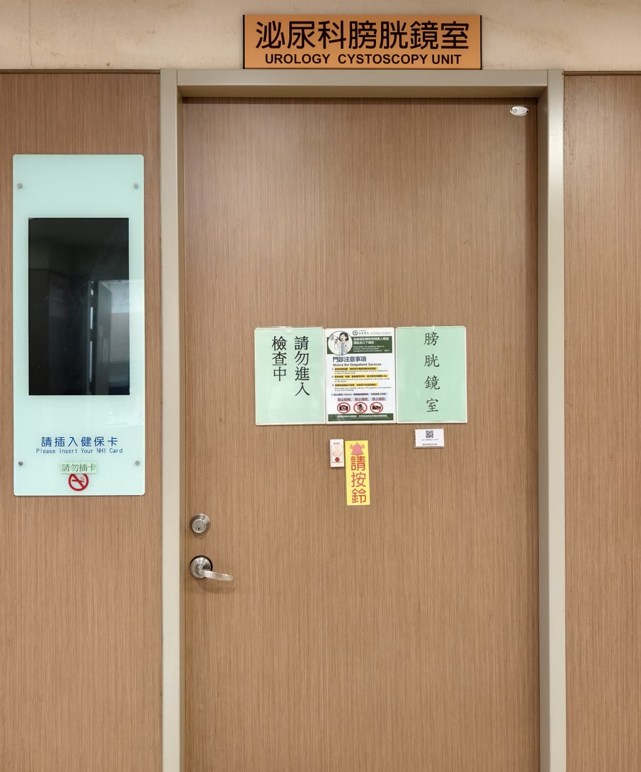 醫院走廊上一扇木紋門，門上方掛著「泌尿科膀胱鏡室 UROLOGY CYSTOSCOPY UNIT」的橘色門牌，門上貼著「請勿進入 檢查中」「膀胱鏡室」的告示和「請按鈴」的標示，左邊牆上是插健保卡的面板