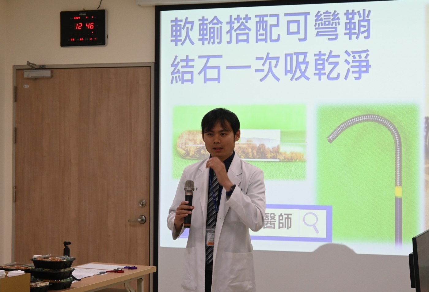 黃韋銘醫師身著白袍手持麥克風站在投影幕旁，幕上寫著「軟輸搭配可彎鞘　結石一次吸乾淨」，旁邊並列著結石檢體與可彎吸石鞘的照片